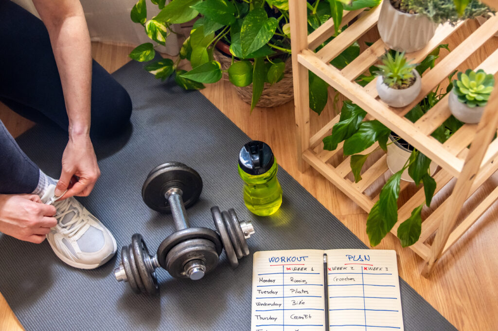 Woman tying her running shoes, preparing for a home workout session on a fitness mat with dumbbells and a workout plan, surrounded by houseplants promoting wellness and a healthy lifestyle