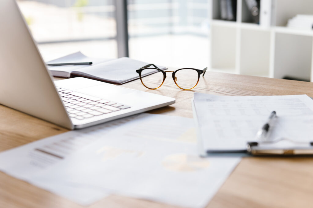 Image of workplace in office room without people with open laptop, clipboard, glasses pencil and notebook lying on wooden desk