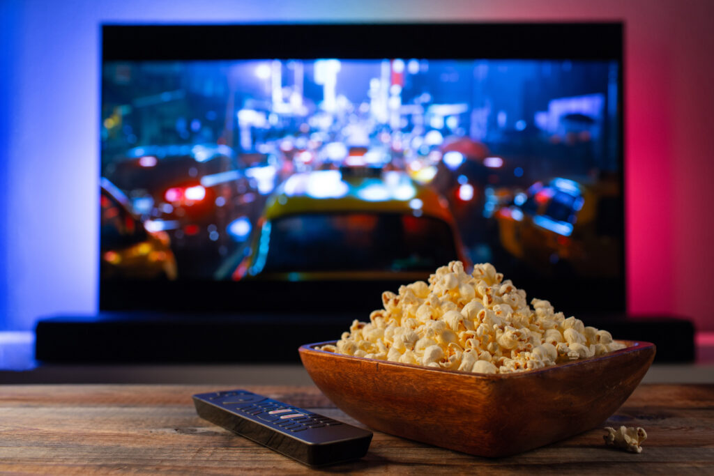 A wooden bowl of popcorn and remote control in the background the TV works. Evening cozy watching a movie or TV series at home.