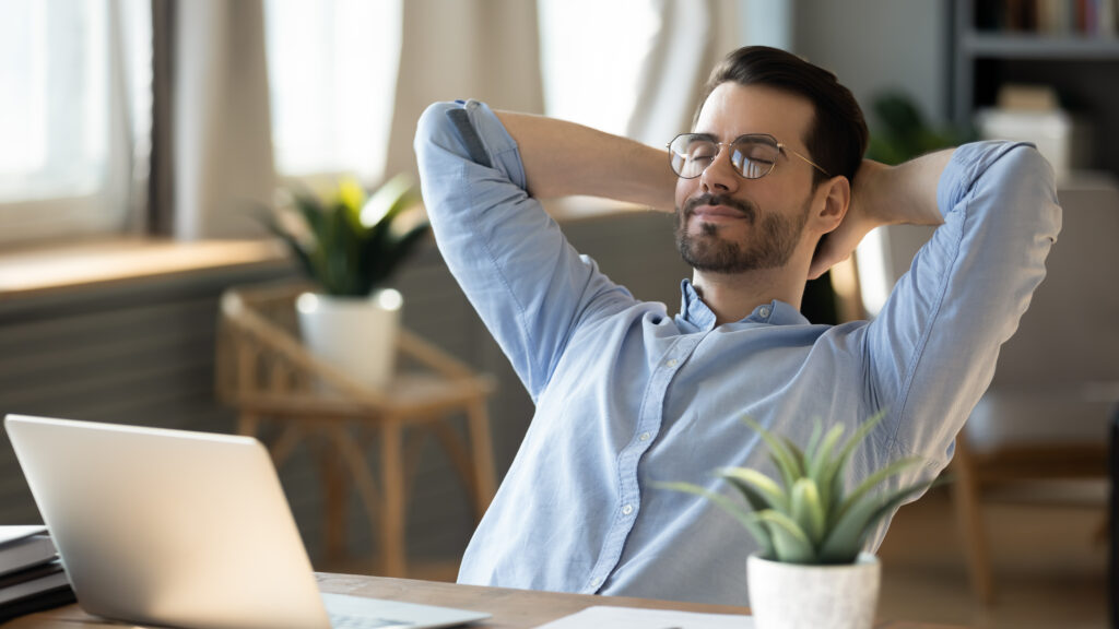 Calm millennial man in glaAsses sit relax at home office workplace take nap or daydream. Happy relaxed Caucasian young male rest in chair distracted from computer work, relieve negative emotions.