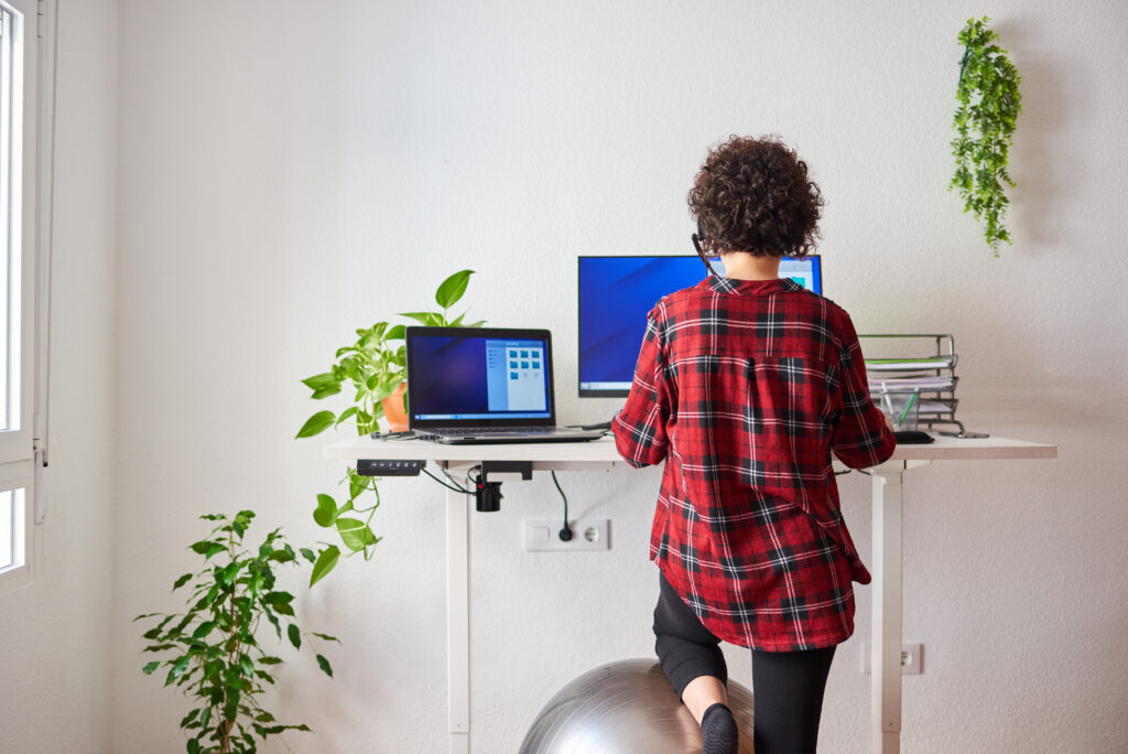 Unrecognizable woman teleworking at an adjustable standing desk with one knee resting on a fitball