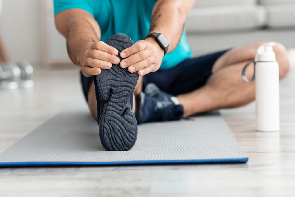 Unrecognizable millennial muscular european man athlete doing leg exercises, stretching on mat with bottle of water in living room interior, close up. Sports training, health care and active lifestyle