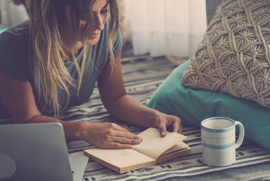 Beautiful young woman reading book while lying on carpet beside laptop and glass at home. Woman spending leisure time lying down on carpet and reading a book at home