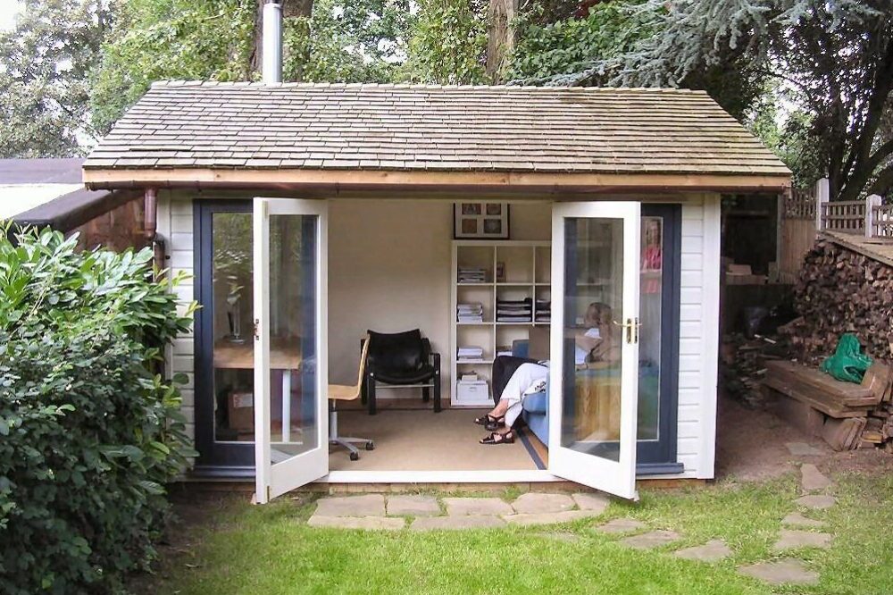 Backyard garden office shed with open double doors, wooden shingle roof, and two people sitting inside.