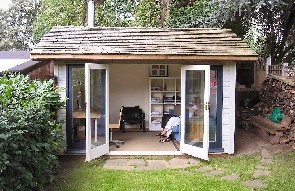 Backyard garden office shed with open double doors, wooden shingle roof, and two people sitting inside.