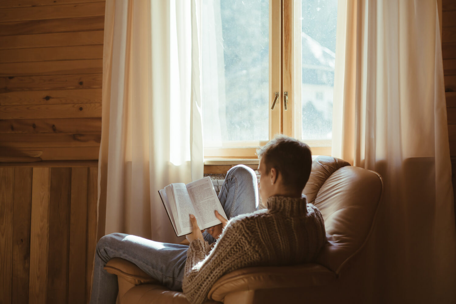 Young man in warm sweater reading book while relaxing on armchair by the window inside cozy log cabin