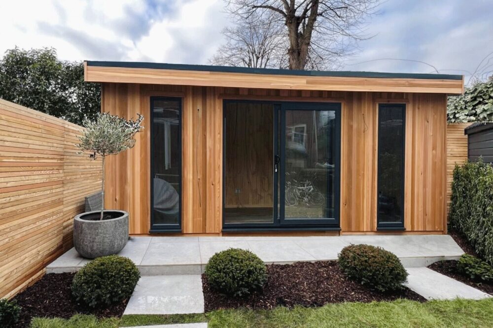Contemporary Garden Shed with Timber Cladding & Glass Doors A contemporary timber-clad garden shed with black framed sliding doors on a cloudy day. Paved patio and landscaped garden.