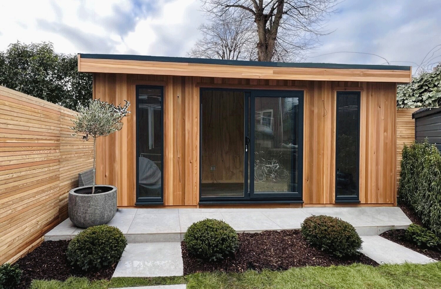 A contemporary timber-clad garden shed with black framed sliding doors on a cloudy day. Paved patio and landscaped garden.