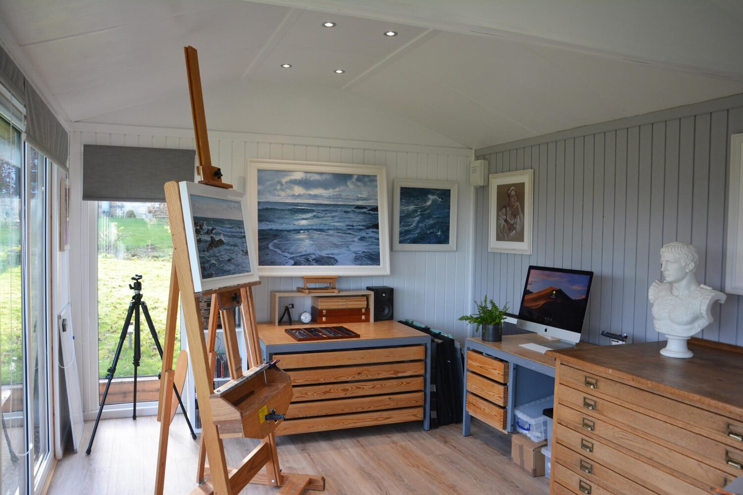 Garden room art studio interior with wooden easel, seascape paintings, desk with a large window overlooking the garden.
