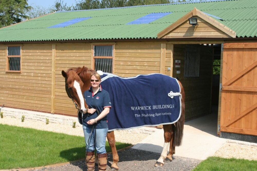 Woman standing with brown horse in front of wooden stables