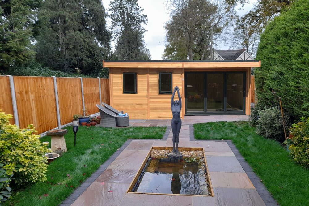 Insulated cedar garden office pod in a UK garden with black-framed sliding glass doors, a window, and a statue with a small pond in the garden exterior.