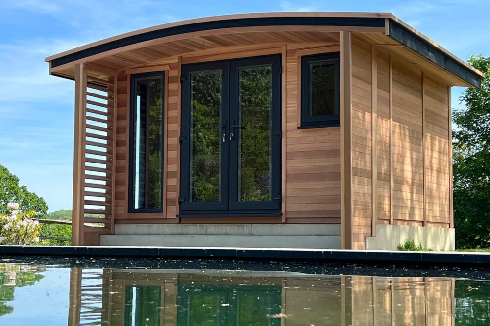 Luxury curved roof garden room featuring natural timber cladding, black-framed glazed double doors, side windows and a covered veranda overlooking a garden pond.