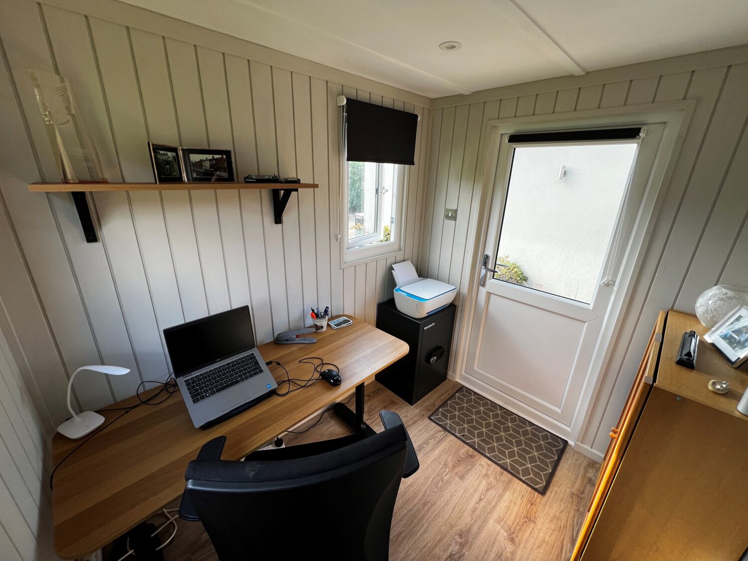 Modern garden office pod interior featuring a wooden desk, laptop, office chair, vertical timber wall panelling, shelving and a glazed entrance door.