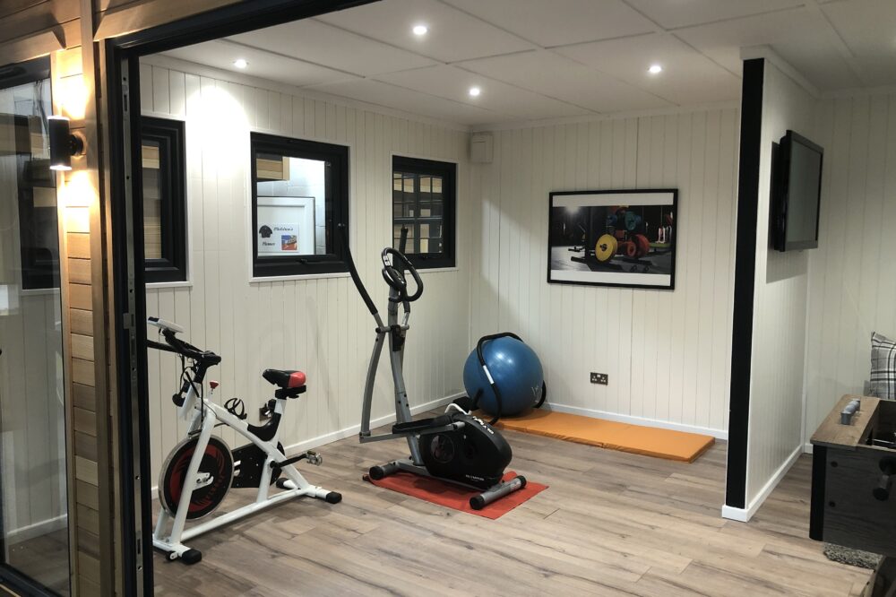Modern garden room home gym with exercise bike, cross trainer, yoga mat, and stability ball. Clean panelled walls and wood flooring.
