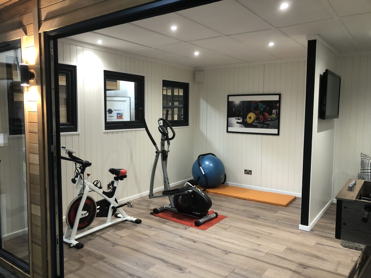 Modern garden room home gym with exercise bike, cross trainer, yoga mat, and stability ball. Clean panelled walls and wood flooring.