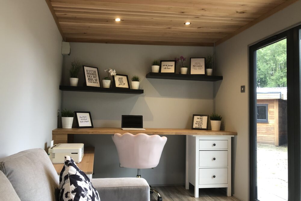 The Elegance garden office pod interior featuring a modern wooden desk, comfortable office chair, timber ceiling, floating shelves and large glass doors.