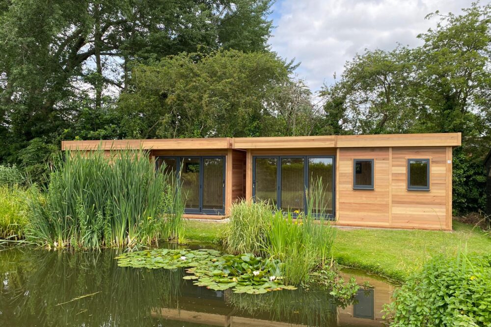 Two adjoining timber garden rooms featuring natural wooden cladding, black-framed glass doors and flat roofs, overlooking a landscaped garden pond.
